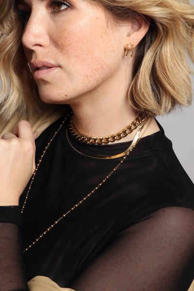 Textured 14k gold plated stainless steel waterproof, non tarnish, water resistant, sustainable chain links by Marrin Costello Jewelry, featuring our geometric double square post hoops, Cuban Link adjustable choker, herringbone 5mm Ramsey Choker, and functional traditional extra long Rachel Rosary in gold, styled with a sheer black turtleneck and khaki vegan leather button down jacket